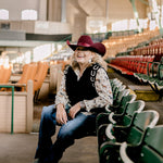 Women wearing a cowboy hat, horse shirt and black sweater vest with horseshoes and sitting in a stadium with green seats.