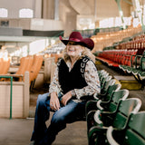 Women wearing a cowboy hat, horse shirt and black sweater vest with horseshoes and sitting in a stadium with green seats.
