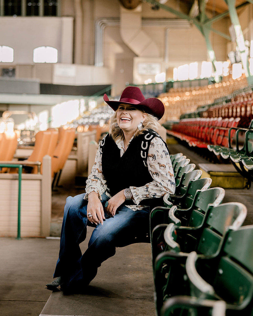 Women wearing a cowboy hat, horse shirt and black sweater vest with horseshoes and sitting in a stadium with green seats.