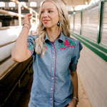 Women wearing a denim pearl snap top with pink western floral embroidery and green leaves. 