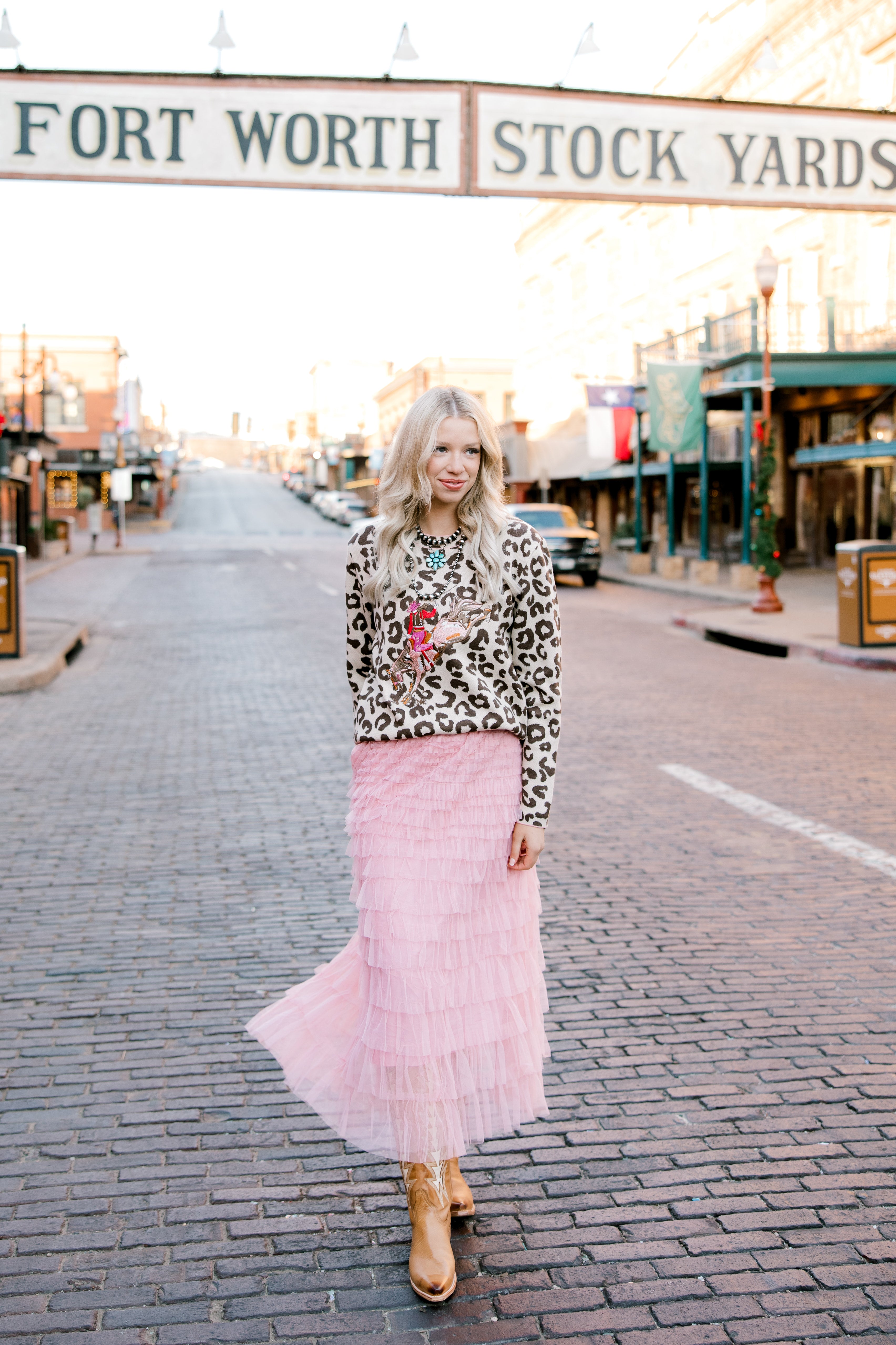Woman in a leopard print top and pink skirt walking on a street with 'Fort Worth Stock Yards' sign in the background.