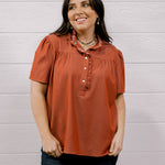 A woman wearing a rust-orange short-sleeve blouse with a ruffled collar and button placket, paired with dark blue jeans, standing against a white wood-paneled wall.