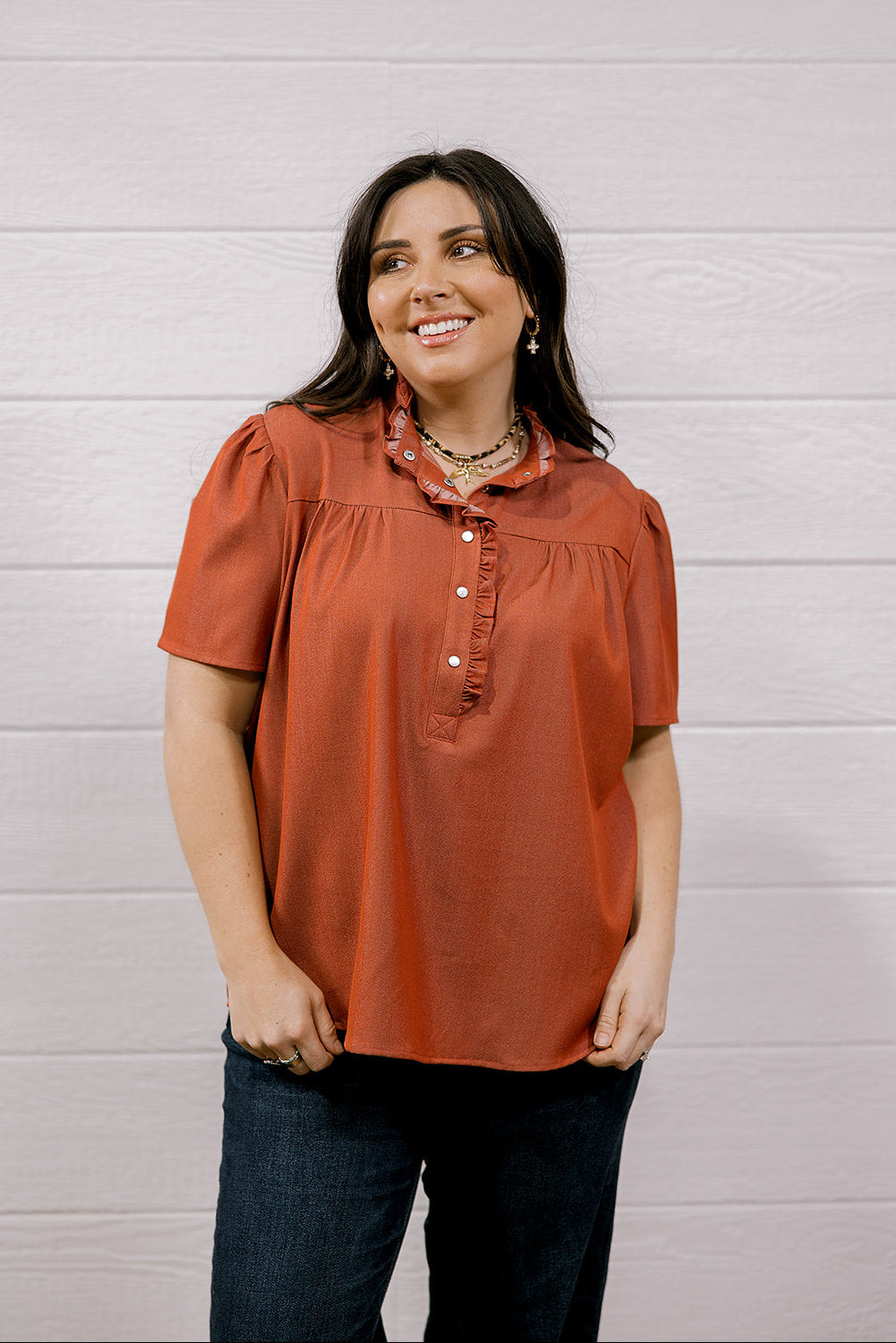 A woman wearing a rust-orange short-sleeve blouse with a ruffled collar and button placket, paired with dark blue jeans, standing against a white wood-paneled wall.