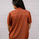 A woman viewed from the back wearing a rust-orange short-sleeve blouse with a gathered neckline and dark pants, standing against a white paneled wall.