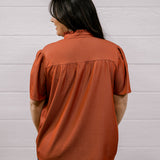 A woman viewed from the back wearing a rust-orange short-sleeve blouse with a gathered neckline and dark pants, standing against a white paneled wall.