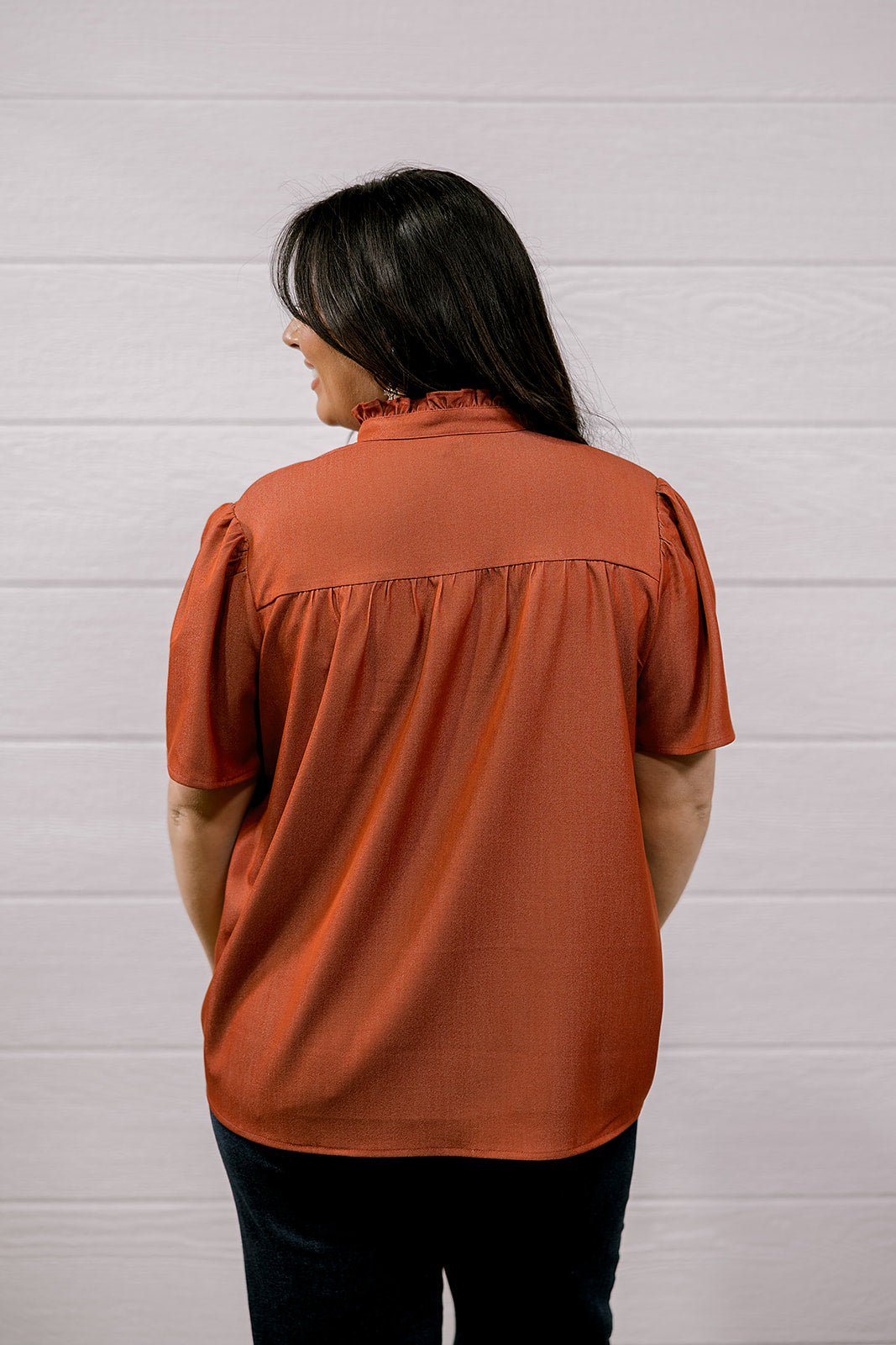 A woman viewed from the back wearing a rust-orange short-sleeve blouse with a gathered neckline and dark pants, standing against a white paneled wall.