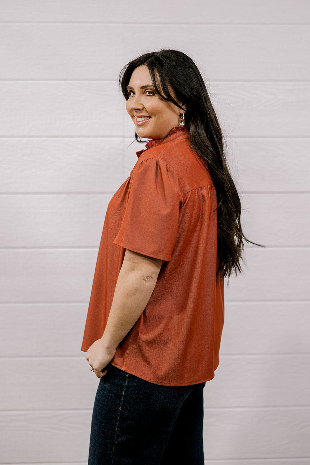 A woman with long dark hair is shown from the side/back wearing a rust-orange short-sleeve blouse and dark jeans, standing against a white paneled wall.