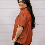 A woman with long dark hair is shown from the side/back wearing a rust-orange short-sleeve blouse and dark jeans, standing against a white paneled wall.