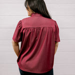 A woman viewed from behind wearing a short-sleeve burgundy blouse with gathered detailing at the back, paired with dark pants, standing against a white paneled wall.