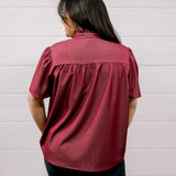 A woman viewed from behind wearing a short-sleeve burgundy blouse with gathered detailing at the back, paired with dark pants, standing against a white paneled wall.