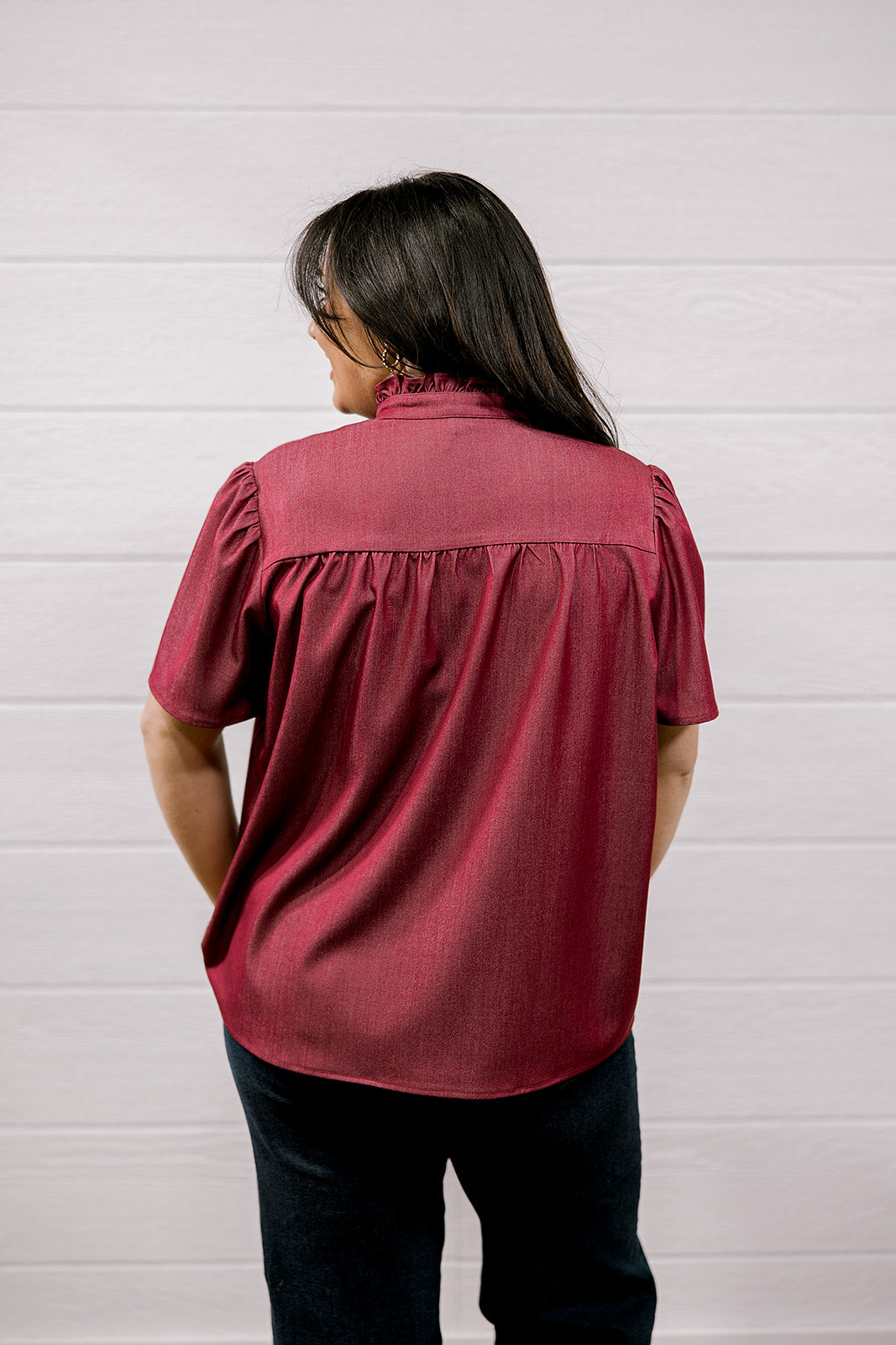 A woman viewed from behind wearing a short-sleeve burgundy blouse with gathered detailing at the back, paired with dark pants, standing against a white paneled wall.