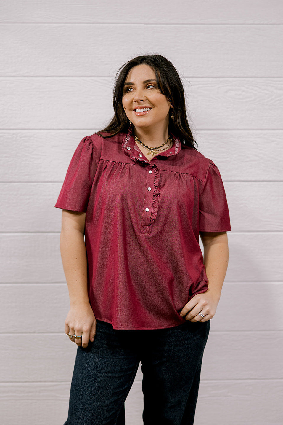 A woman wearing a short-sleeve burgundy button-up blouse with a ruffled collar and dark jeans, standing against a white wood-paneled wall.