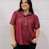 A woman wearing a short-sleeve burgundy button-up blouse with a ruffled collar and dark jeans, standing against a white wood-paneled wall.