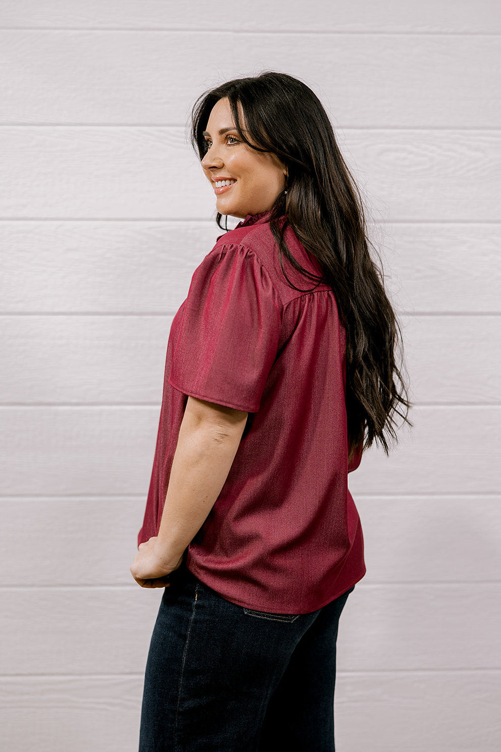 A woman with long dark hair is shown from the side and back, wearing a short-sleeve maroon blouse and dark jeans, standing against a white wood-paneled wall.