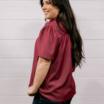 A woman with long dark hair is shown from the side and back, wearing a short-sleeve maroon blouse and dark jeans, standing against a white wood-paneled wall.