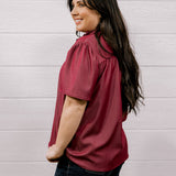A woman with long dark hair is shown from the side and back, wearing a short-sleeve maroon blouse and dark jeans, standing against a white wood-paneled wall.
