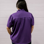 A person viewed from behind wearing a purple short-sleeve blouse and black pants, standing against a white paneled wall.