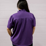 A person viewed from behind wearing a purple short-sleeve blouse and black pants, standing against a white paneled wall.