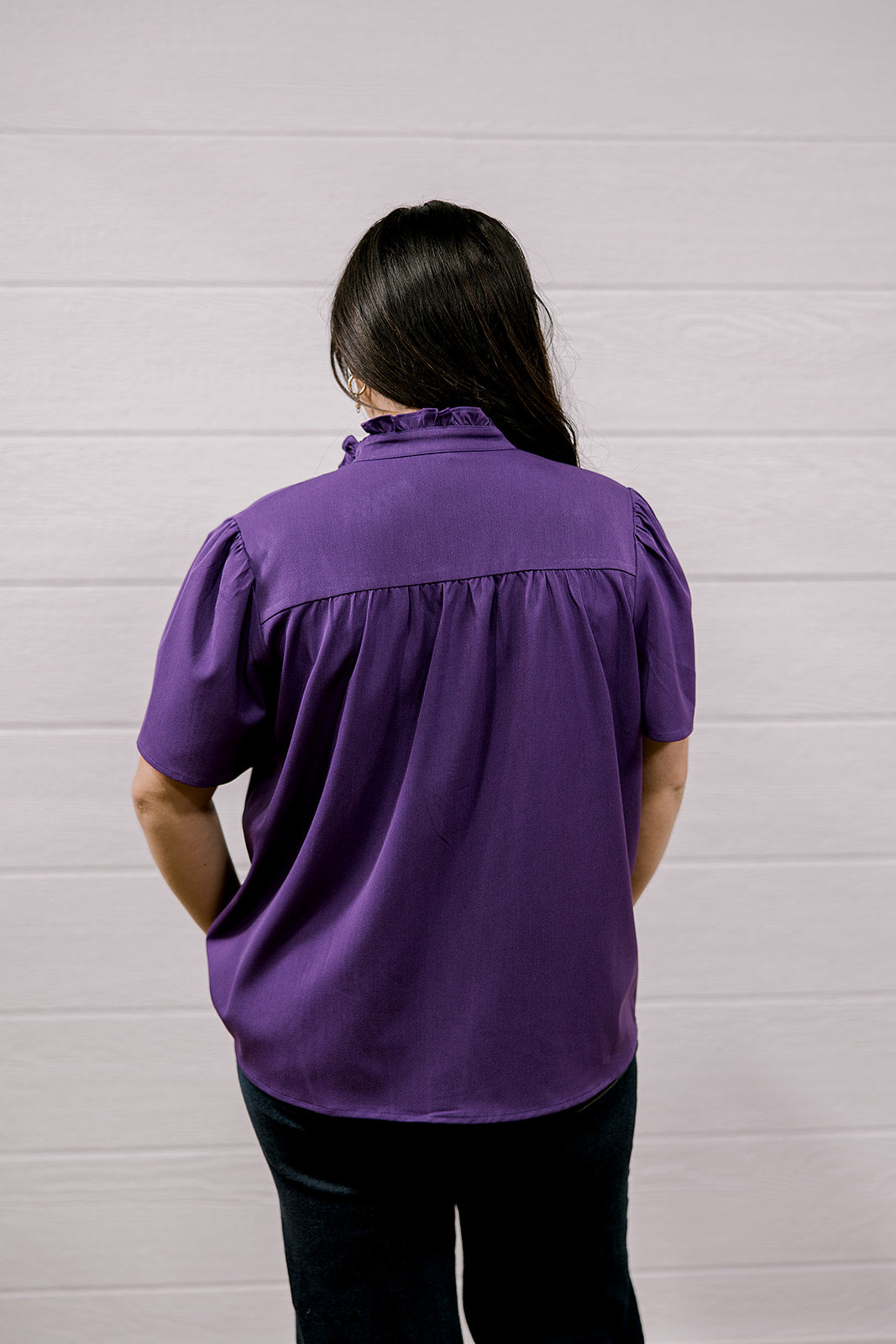 A person viewed from behind wearing a purple short-sleeve blouse and black pants, standing against a white paneled wall.