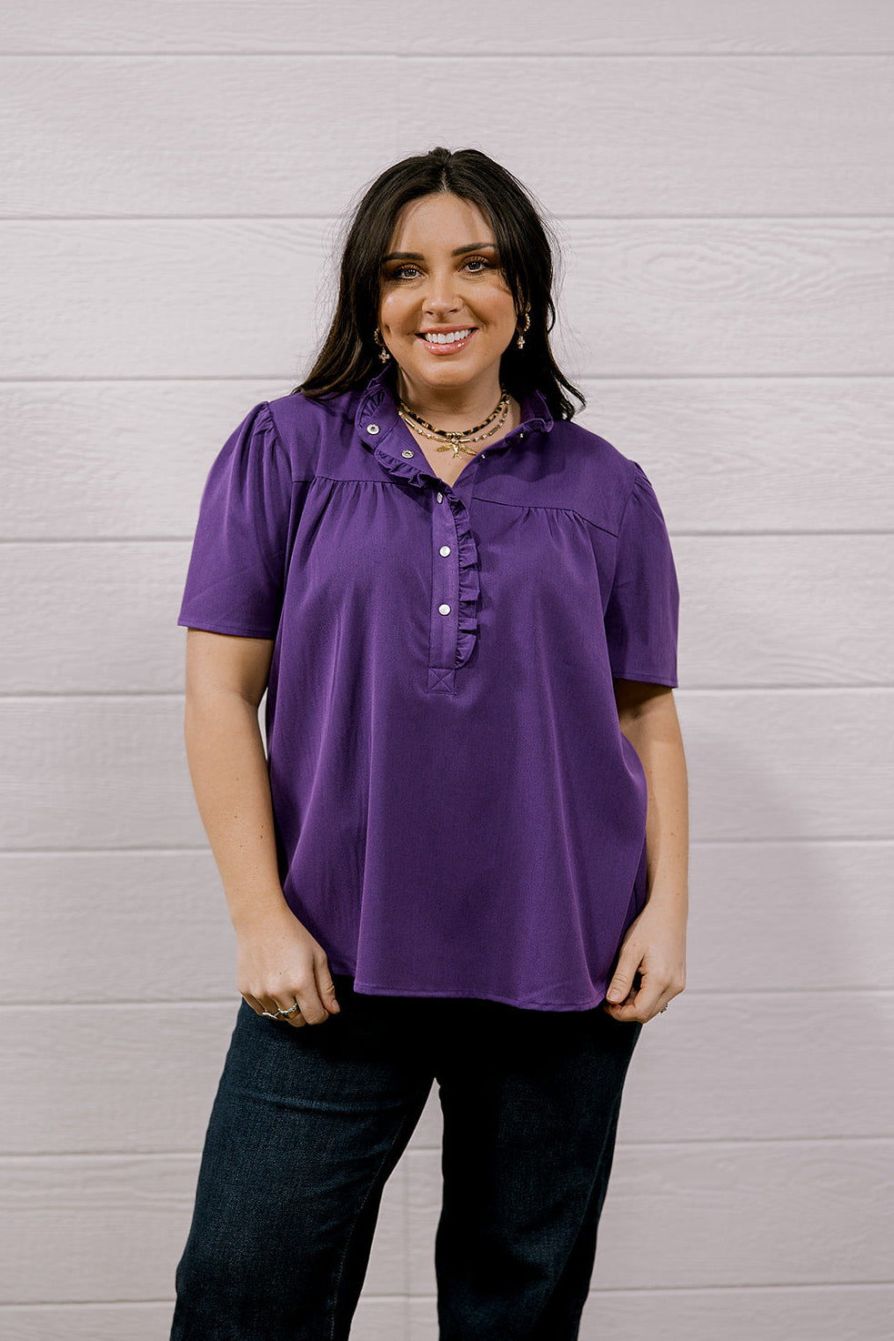 A woman modeling a purple short-sleeve button-up blouse with ruffle details on the collar and chest, paired with dark jeans, standing against a white wood-paneled wall.
