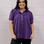 A woman modeling a purple short-sleeve button-up blouse with ruffle details on the collar and chest, paired with dark jeans, standing against a white wood-paneled wall.