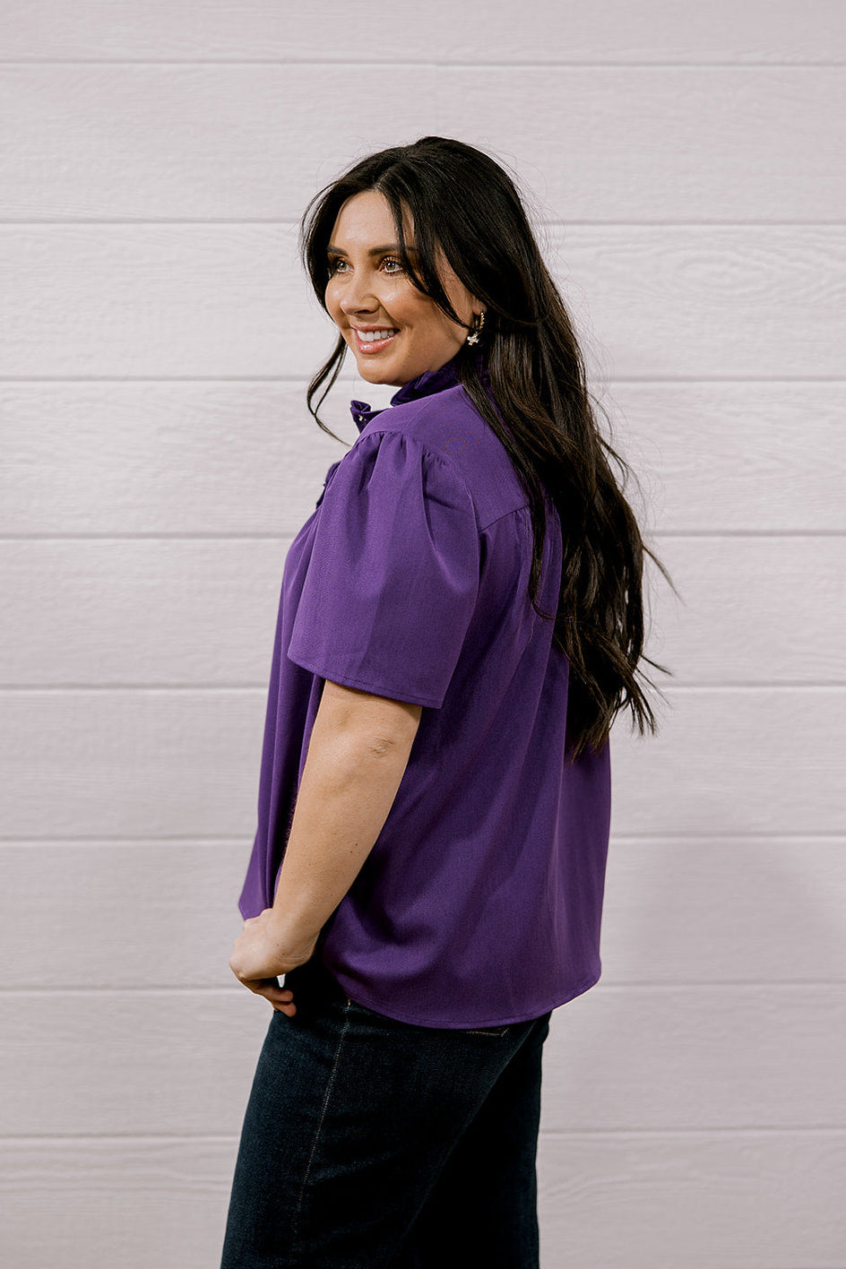 A woman with long dark hair is shown from the back wearing a purple short-sleeve blouse and dark jeans, standing against a white wood-paneled wall.