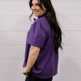 A woman with long dark hair is shown from the back wearing a purple short-sleeve blouse and dark jeans, standing against a white wood-paneled wall.