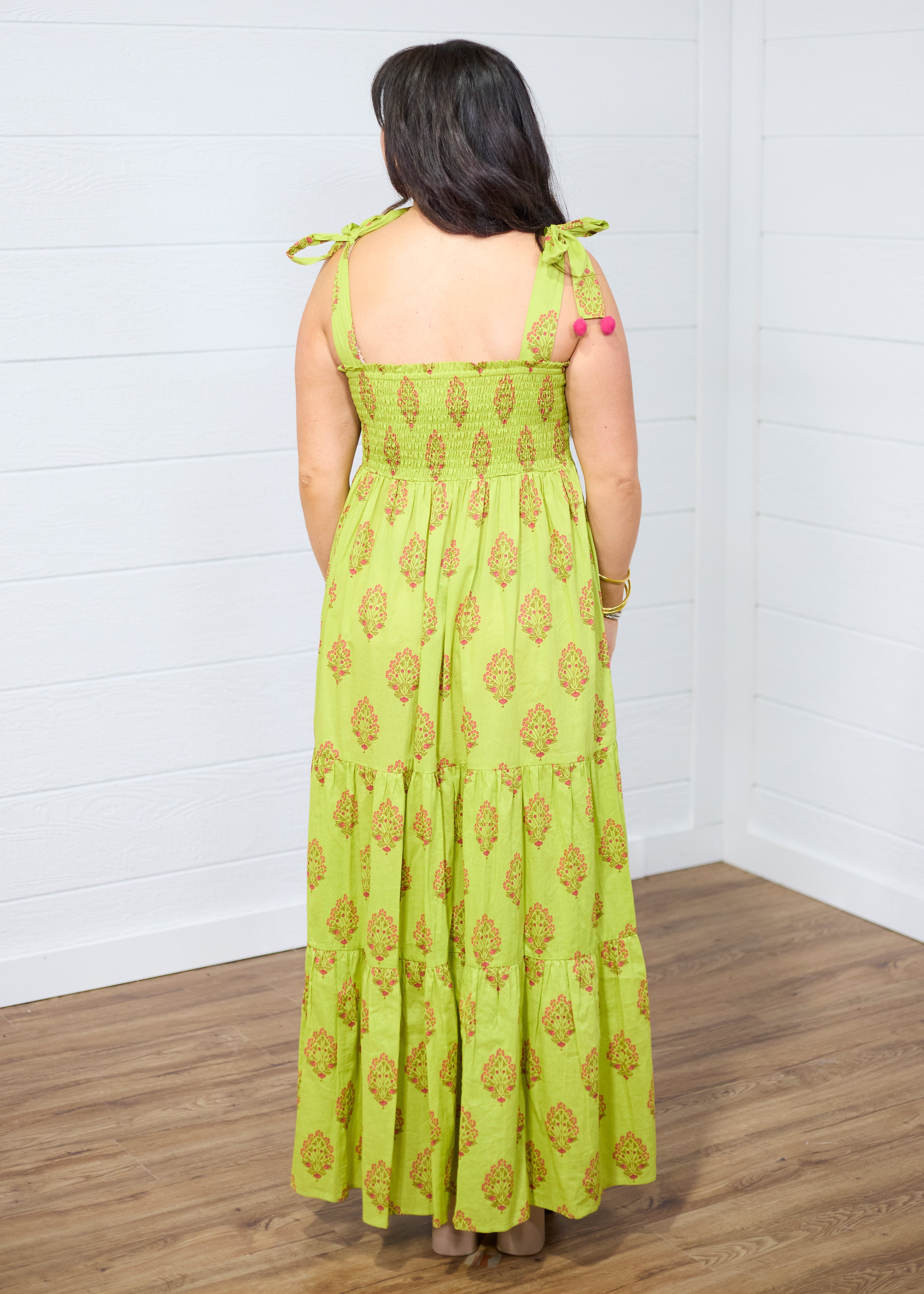 Woman with dark hair standing in a back profile position wearing the Addison Maxi Dress against a white background. The image focal point is of the back of the dress showing off the graphics and sleeveless top. 