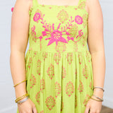 Woman with dark hair standing in a front profile position with a deep focal length towards the top embroidery on dress. This shows the floral print and the pom-poms on tied straps. 