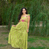 Woman with dark hair in a lifestyle scene wearing the Addison Maxi Dress. The image background is heavy with greenery. Model has paired dress with white sandals.