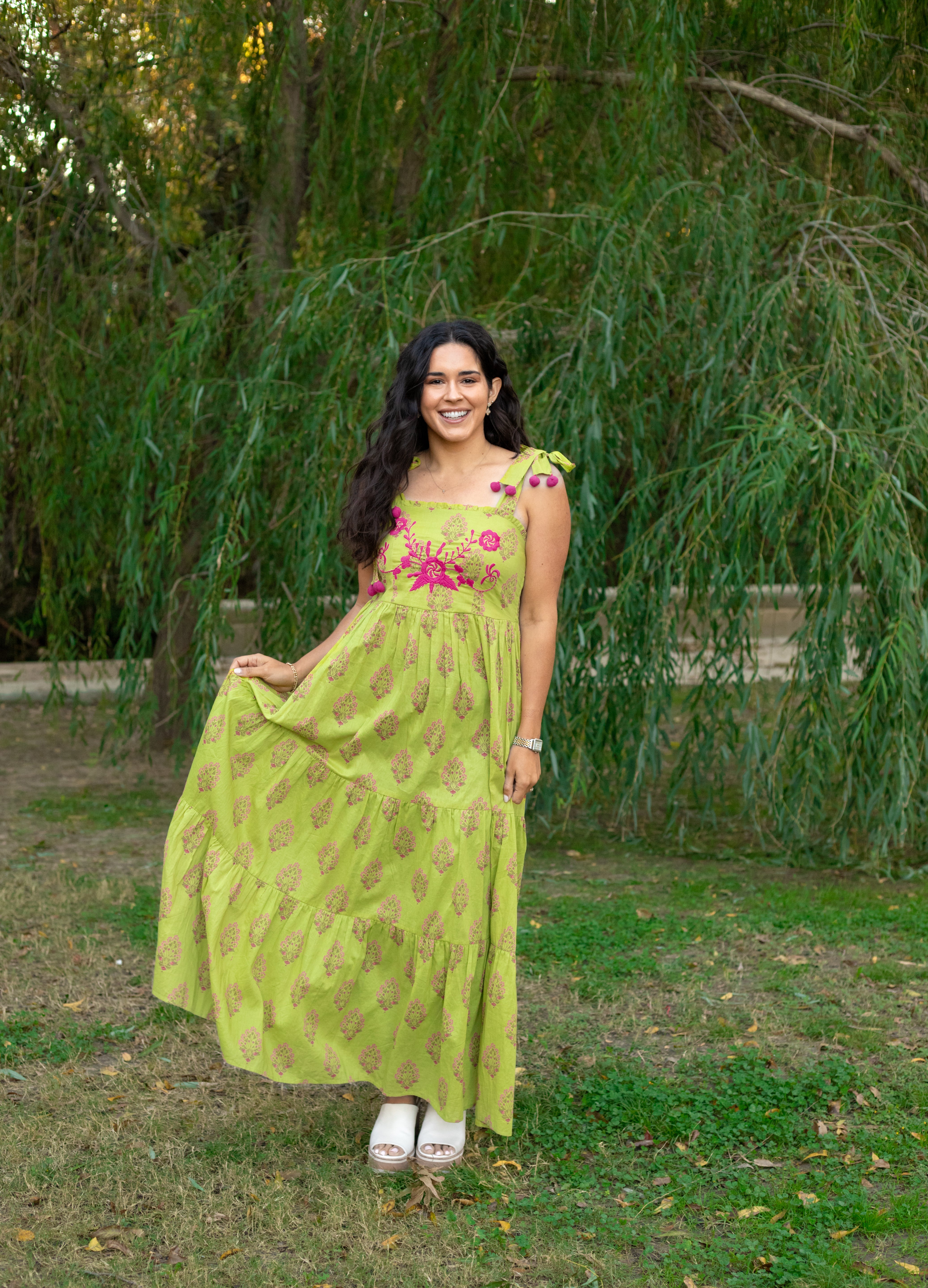 Woman with dark hair in a lifestyle scene wearing the Addison Maxi Dress. The image background is heavy with greenery. Model has paired dress with white sandals.
