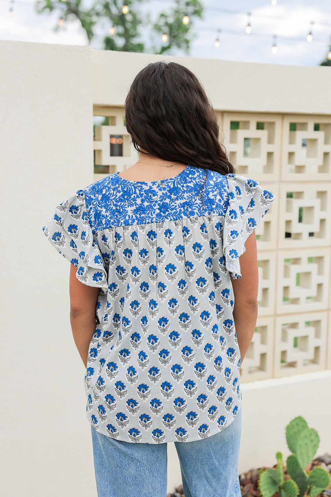 Woman with dark hair standing in a back profile position wearing the Alura Top in a lifestyle scene. The focal point of this image is the blue floral and blue embroidery. The model paired this top with light-wash denim jeans. 