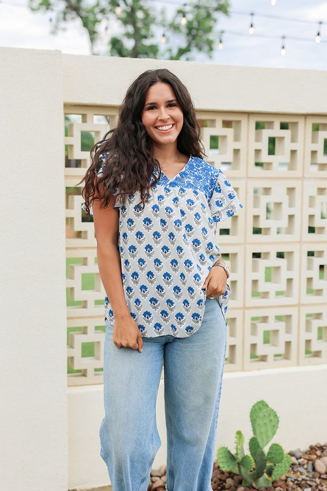 Woman with dark hair standing in a front profile position wearing the Alura Top in a lifestyle scene. The focal point of this image is the floral pattern and blue embroidery across chest with v-neckline. The model paired this top with light-wash denim jeans. 