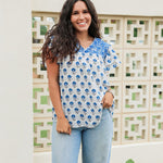 Woman with dark hair standing in a front profile position wearing the Alura Top in a lifestyle scene. The focal point of this image is the floral pattern and blue embroidery across chest with v-neckline. The model paired this top with light-wash denim jeans. 