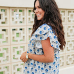 Woman with dark hair standing in a side profile position wearing the Alura Top in a lifestyle scene. The focal point of this image is the ruffled sleeves and floral print. The model paired this top with light-wash denim jeans. 