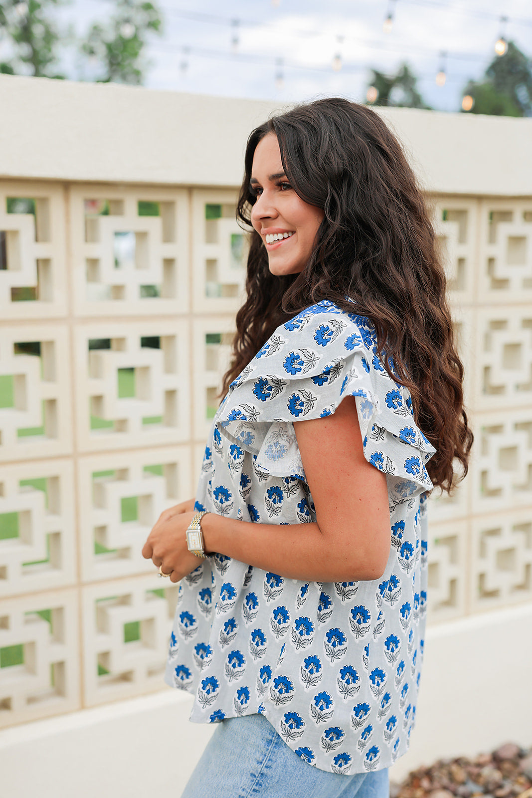 Woman with dark hair standing in a side profile position wearing the Alura Top in a lifestyle scene. The focal point of this image is the ruffled sleeves and floral print. The model paired this top with light-wash denim jeans. 