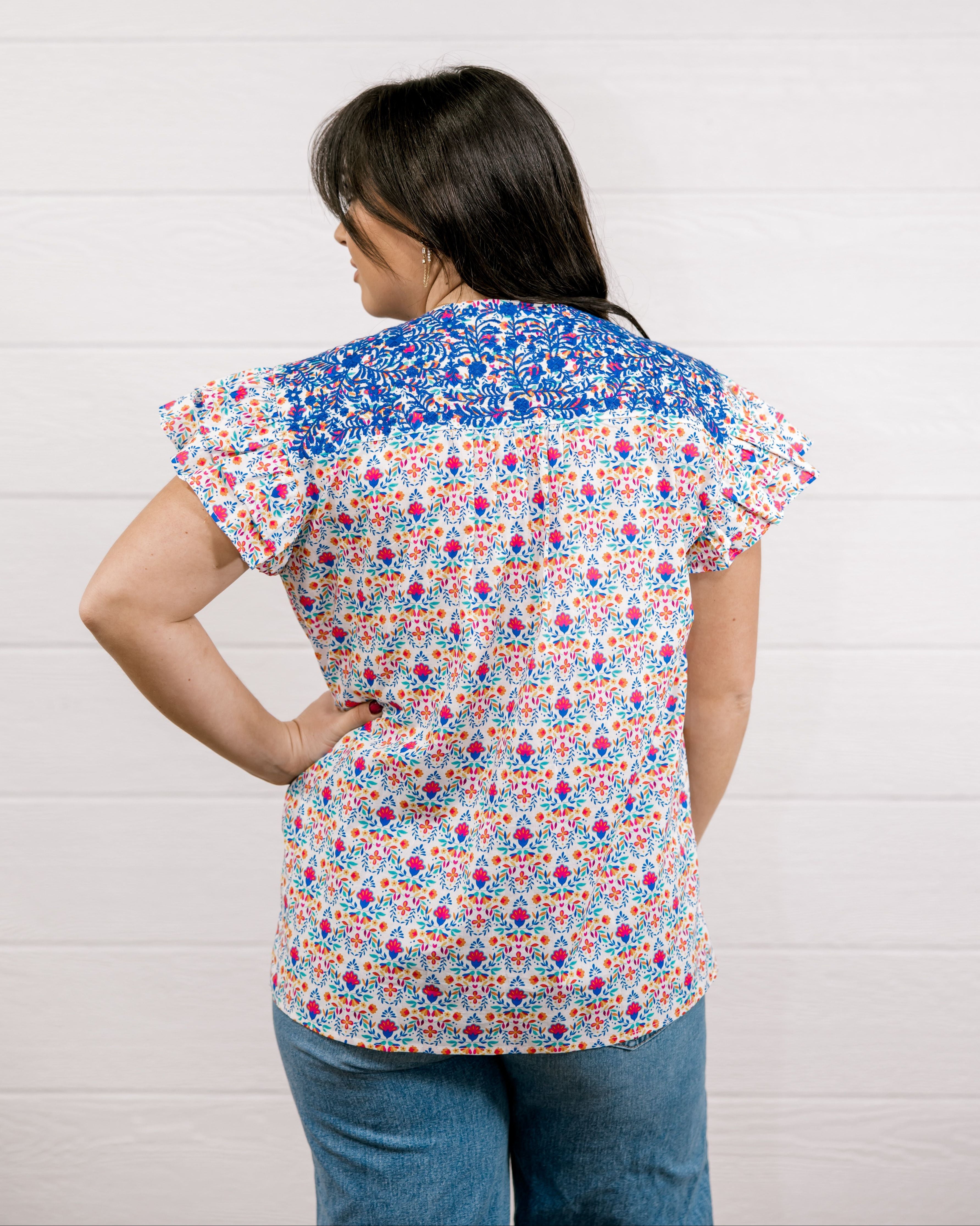 Women wearing a colorful top with blue ebroidery on the top showing the back on a white background 