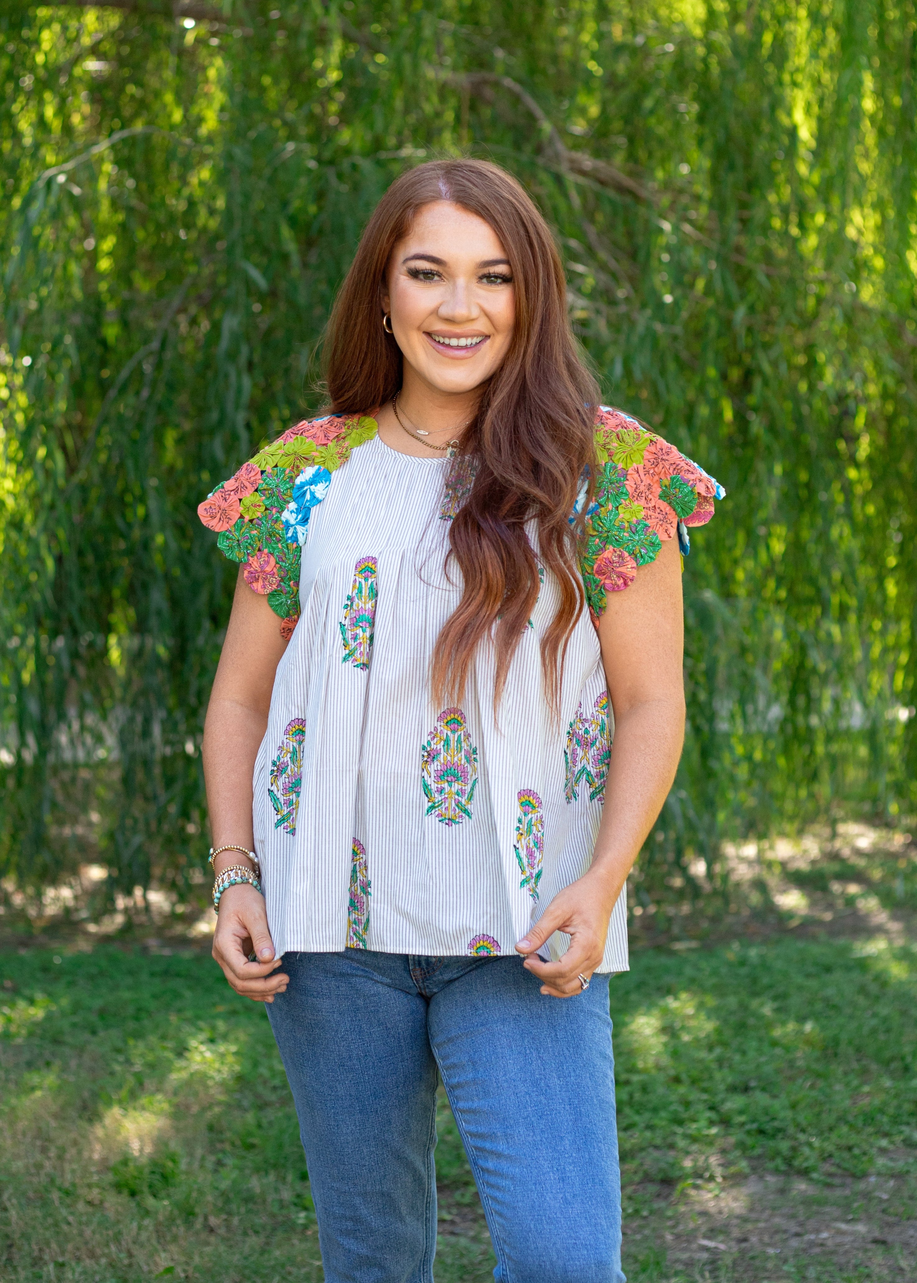 Woman with red hair standing in a front facing position in a lifestyle scene. The back ground shows a lot of greenery, and woman hair top paired medium-wash jeans.