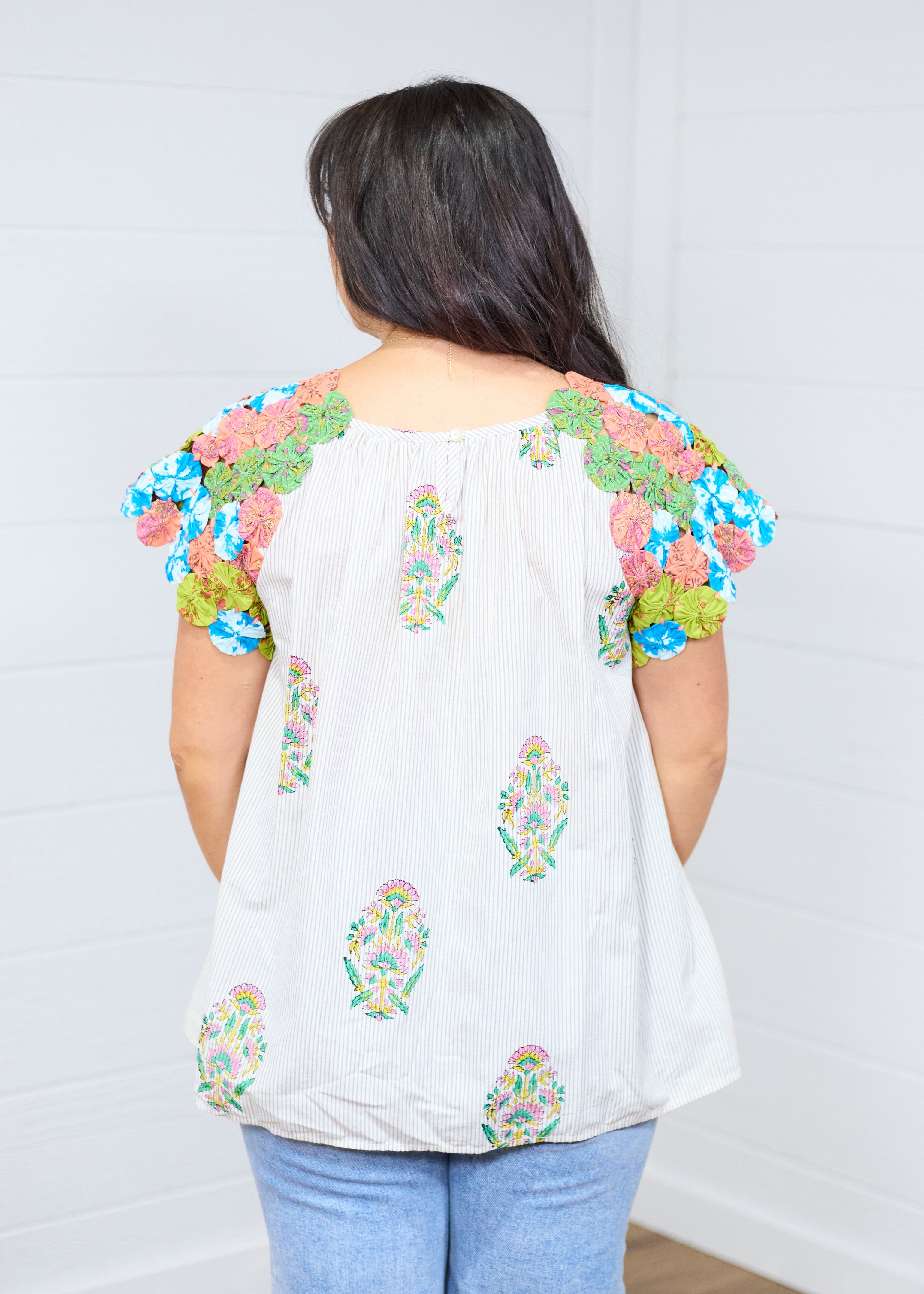 Woman with dark brown hair standing in back profile to show off back of the Asher Top. She is against a white background, and paired the top with light-wash denim jeans. 