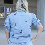 Woman with blonde hair standing in a  back profile position wearing the Bailey Top in a lifestyle scene. The focal point of this image is the purple embroidered boots on back of top as well as back of ruffled sleeves. The model paired this top with light-wash denim shorts. 