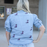 Woman with blonde hair standing in a  back profile position wearing the Bailey Top in a lifestyle scene. The focal point of this image is the purple embroidered boots on back of top as well as back of ruffled sleeves. The model paired this top with light-wash denim shorts. 
