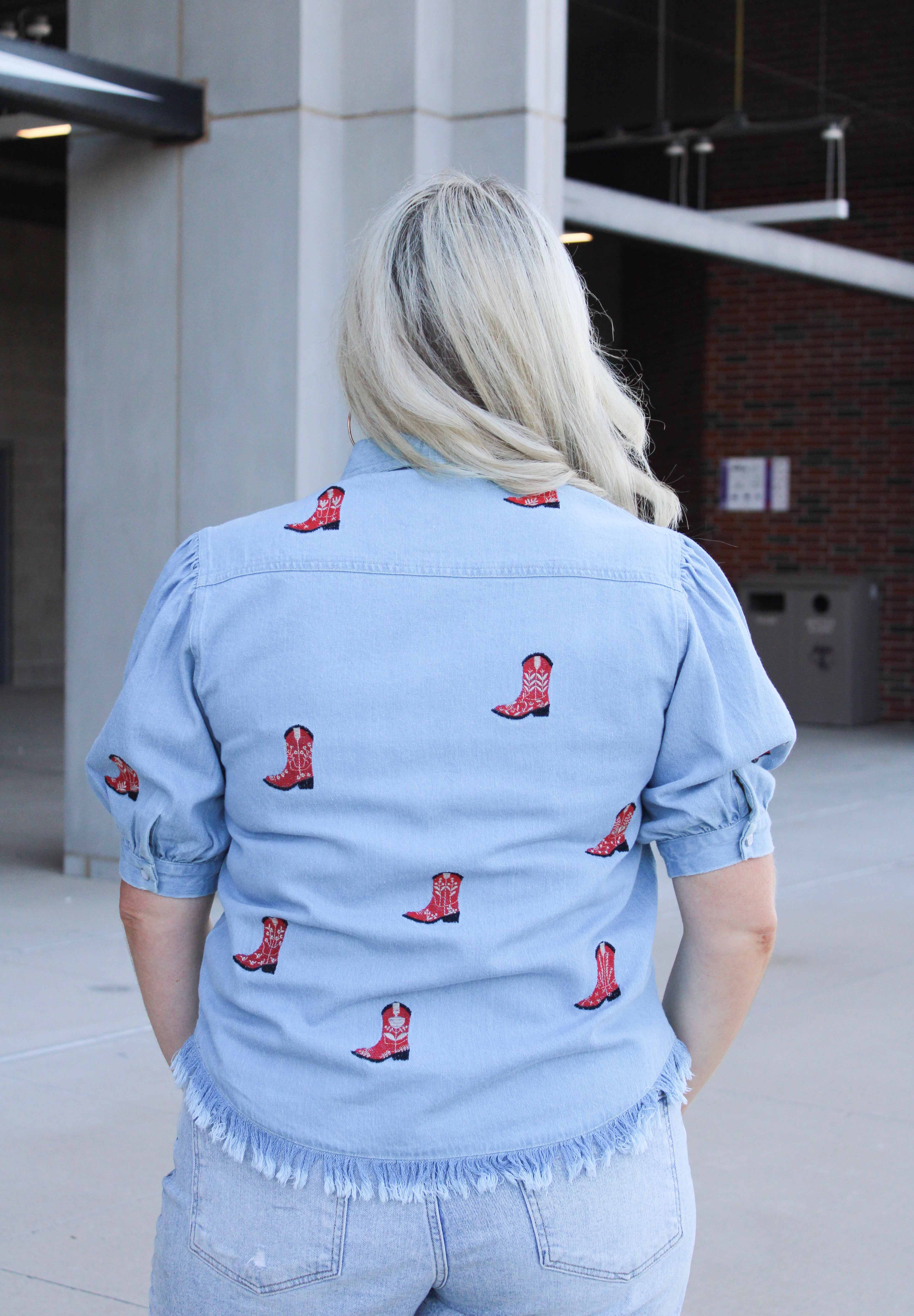 Woman with blonde hair standing in a back profile position wearing a short-sleeve denim top with red embroidered boots. The model paired this top with light-wash denim jeans. 