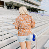 Woman with blonde hair standing in a back profile position wearing the Bell Top at a football field. The focal point of this image is the floral graphics on back and shoulders as well as the ruffled sleeves. The model paired this top with light-wash denim shorts.