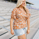 Woman with blonde hair standing in a side profile position wearing the Bell Top at a football field. The focal point of this image is the ruffled sleeves with emphasis on tassels attached to collar and length of top. The model paired this top with light-wash denim shorts and brown cowboy boots.