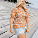 Woman with blonde hair standing in a side profile position wearing the Bell Top at a football field. The focal point of this image is the ruffled sleeves with emphasis on tassels attached to collar and length of top. The model paired this top with light-wash denim shorts and brown cowboy boots.