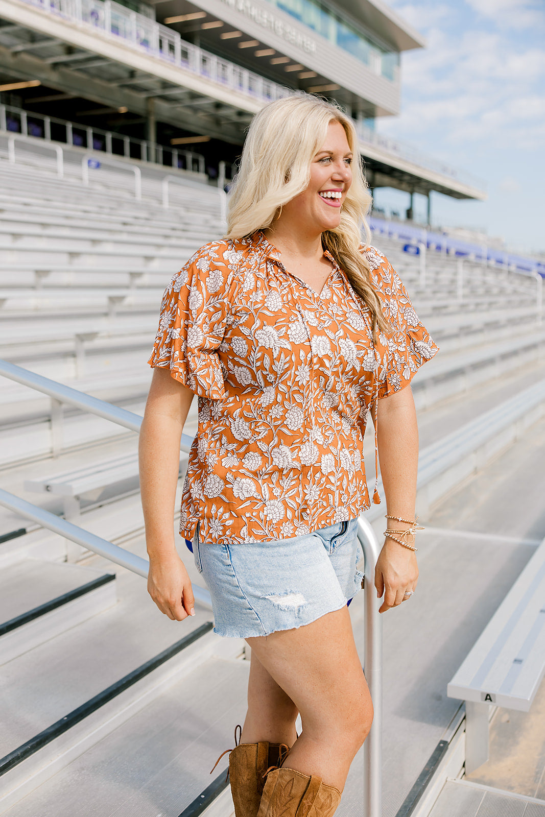 Woman with blonde hair standing in a side profile position wearing the Bell Top at a football field. The focal point of this image is the ruffled sleeves with emphasis on tassels attached to collar and length of top. The model paired this top with light-wash denim shorts and brown cowboy boots.
