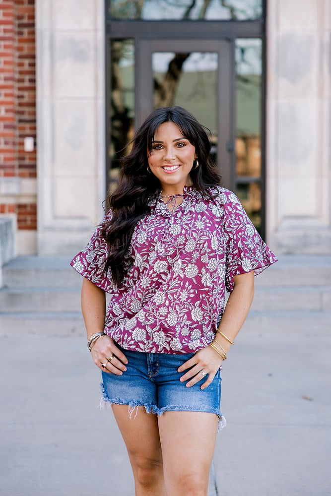 Woman with dark hair wearing short sleeve top with floral graphics and dark-wash denim shorts. 
