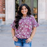 Woman with dark hair wearing short sleeve top with floral graphics and dark-wash denim shorts. 