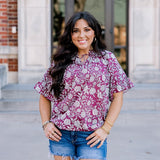 Woman with dark hair wearing short sleeve top with floral graphics and dark-wash denim shorts. 