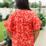 Woman with dark hair standing in a back profile position wearing the Bell Top in a lifestyle scene. The focal point of this image is the white floral print against a red background, ruffled sleeves and pleating on collar. The model paired this top with light-wash denim jeans. 
